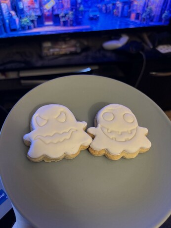 Two ghost-shaped cookies decorated with smooth white royal icing sit on a light grey plate. The cookie on the left has a spooky face with sharp eyes and a jagged mouth, while the cookie on the right has a cheerful face with round eyes and a big smiling mouth with small teeth.