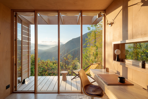 A photograph showing Hermitage Cabin, a small wooden house in the Italian Apennines designed as a modern chalet with a Zen aesthetic. The exterior is clad in warm timber, with a steep gabled roof and broad glass doors opening to mountain views. Inside, every surface is wood — floors, walls, ceiling — giving the room a golden glow. Large windows frame the surrounding forest and rolling peaks, filling the minimalist interior with shifting natural light. The cabin is compact yet open, a balance of simplicity and warmth, appearing at once handcrafted and architectural. It sits quietly in the landscape, almost disappearing into the trees, as if designed for meditation and retreat.
Via Archilovers.com