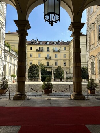 A picturesque courtyard view framed by archways and columns. A red carpet leads toward the open space, showcasing greenery and decorative planters. The background features yellow buildings with balconies, under a clear blue sky.