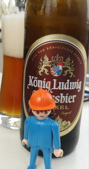 Playmobil figure with orange hard hat standing in front of a beer bottle labeled "König Ludwig Weißbier Dunkel"