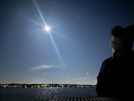 Nighttime photo of the moon over a lake. Moonlight glistens on the water’s surface. City lights are visible on the opposite shore. In the foreground is the silhouette of a woman looking over the lake.
