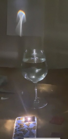 Tape is placed over a cell phone flashlight to form a slit. The phone is placed on a table with the light shining up under a red wine glass filled with water. The light from the phone is refracted and forms a rainbow on a nearby wall.
