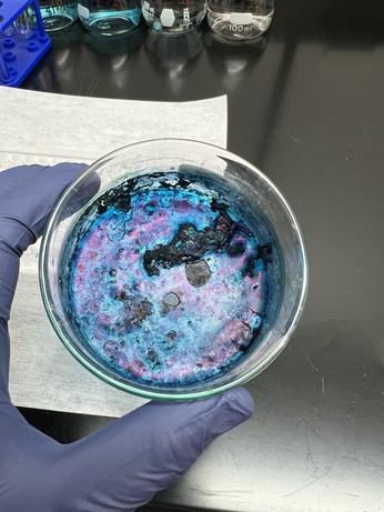 Class bowl being held by a gloved hand. The bowl bottom is covered with an iridescent blue and purple coating, along with some dark blue lumps.