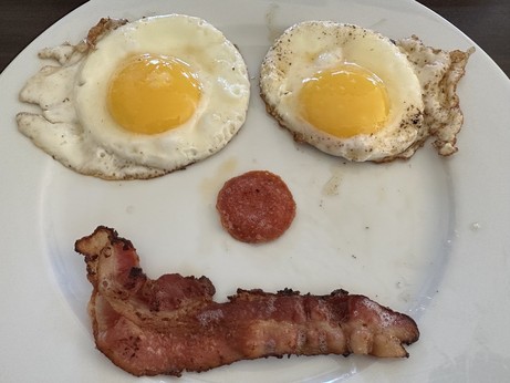 Two eggs are eyes, a pepperoni slice is a nose, and a strip of bacon is a crooked smile, making a face on a white plate. 