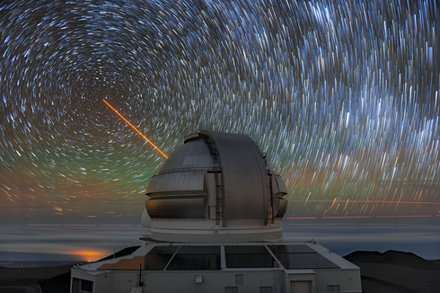 A time lapse image of a test of Gemini North's new circumpolar laser. The image shows the observatory in the foreground, circular star trails, and an orange laser aimed at the sky.