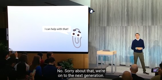 Still from the announcement of Microsoft's “AI-powered Bing.”  Clippy is shown on the projection screen with a balloon reading “I can help with that!”  Microsoft manager Yusuf Mehdi comments: “No.  Sorry about that, we're on to the next generation.”  <https://youtu.be/rOeRWRJ16yY?t=868>