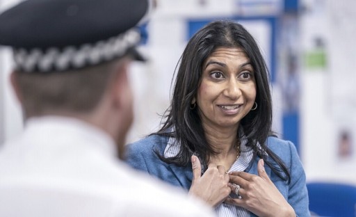 Suella Braverman with a gormless grinning expression holding up her hands in what appears to be a “thumbs up” pose, faced by one or more police officers, reminiscent of the infamous sarcastic “friend!” scene from The Inbetweeners. 