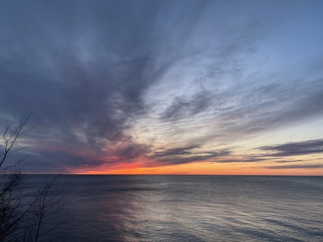 Dramatic grey clouds emanating up from an Orange sunset over Lake Superior. Reflecting in the water, dark other left and lighter on the right. 