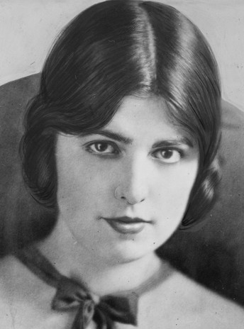 black and white photo of Virginia Rappe, she has dark hair parted in the middle, pulled back, she is wearing a ribbon/bow around her neck, she is looking directly at the camera