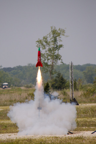 A short, 4" diameter, red rocket that looks like a Sriracha bottle leaves the pad on a flame as long as the rocket with a cloud of gray smoke below it.