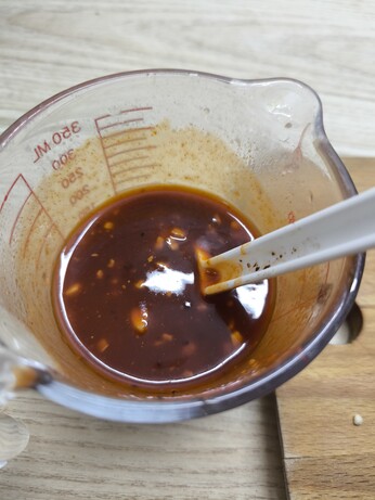 A small pyrex jug is shown, top down view. There is a white plastic spoon in the sauce. The sauce is a deep, smoky red with chunks of garlic and chilli flakes visible.